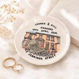 May include: A round white ceramic trinket dish featuring a hand-painted illustration of a brick house. The text reads LAUREN & BILL ENGAGED 12TH APRIL 2025 and 12 JENNINGS STREET. Two gold-toned metal rings are placed next to the dish on a white fabric.
