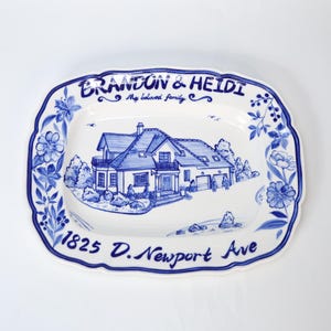 May include: A white ceramic serving platter with a blue floral border and a hand-painted illustration of a house. The platter is inscribed with the names "Brandon & Heidi", the phrase "My beloved family", and the address "1825 D. Newport Ave".