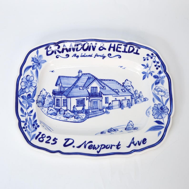 May include: A white ceramic serving platter with a blue floral border and a hand-painted illustration of a house. The platter is inscribed with the names "Brandon & Heidi", the phrase "My beloved family", and the address "1825 D. Newport Ave".