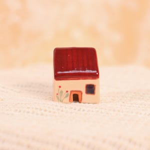 May include: A miniature ceramic house with a red roof, beige walls, and a small square window. The house features a brown door and a painted floral design. The roof has a textured appearance.