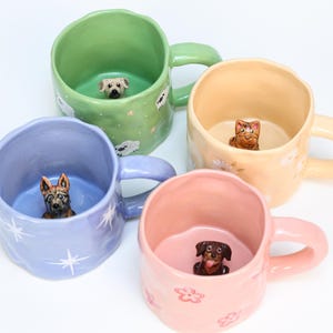 Op de afbeelding: Vier keramische mokken in pastelkleuren: groen, geel, lavendel en roze. Elke mok heeft een gebeeldhouwd dierenfiguur aan de binnenkant: een hond, een kat en twee verschillende hondenrassen. De mokken hebben oren en zijn versierd met bloemen- en stippenpatronen.