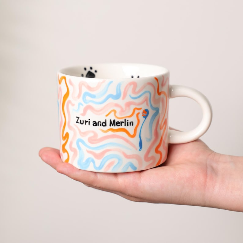 K&ouml;nnte beinhalten: Wei&szlig;e Keramiktasse mit einem skurrilen Design. Die Tasse hat ein Muster aus orangefarbenen, blauen und rosa Wellenlinien mit den Worten "Zuri and Merlin" in Schwarz. Schwarze Pfotenabdr&uuml;cke befinden sich im Inneren des Rands.