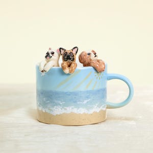 May include: A ceramic mug with a beach scene design, featuring a light blue sky and ocean, sandy shore, and a yellow sun. The mug is adorned with three small, sculpted animal figures: a white cat, a brown and black dog, and a brown dog.