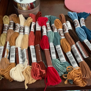 May include: A collection of embroidery floss spools in various colors, including red, orange, yellow, green, blue, brown, and beige. Each spool has a label with a DMC number and the brand name.