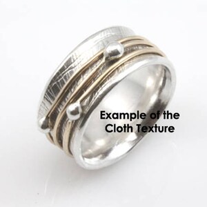 May include: A silver and gold ring with a textured finish and three gold bands. The ring has two small silver beads on the bands. "Example of the Cloth Texture" is written on the ring.
