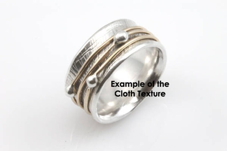 May include: A silver and gold ring with a textured finish and three gold bands. The ring has two small silver beads on the bands. "Example of the Cloth Texture" is written on the ring.