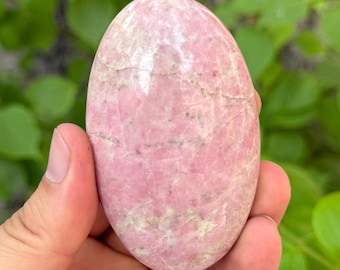 May include: A smooth, oval-shaped pink gemstone with light-colored veining. The polished stone is held in a hand, with a green leafy background. The gemstone's color ranges from light to medium pink.