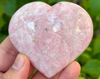Puede incluir: Una piedra en forma de corazón con un patrón veteado rosa y blanco. La piedra se sostiene en una mano, con follaje verde en el fondo. La piedra parece pulida y lisa.