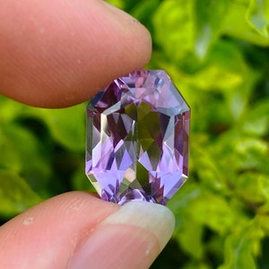 Peut inclure: Une grande gemme d'am&eacute;thyste facet&eacute;e de forme octogonale avec une teinte violette. La gemme est tenue dans une main, avec un fond de feuilles vertes.