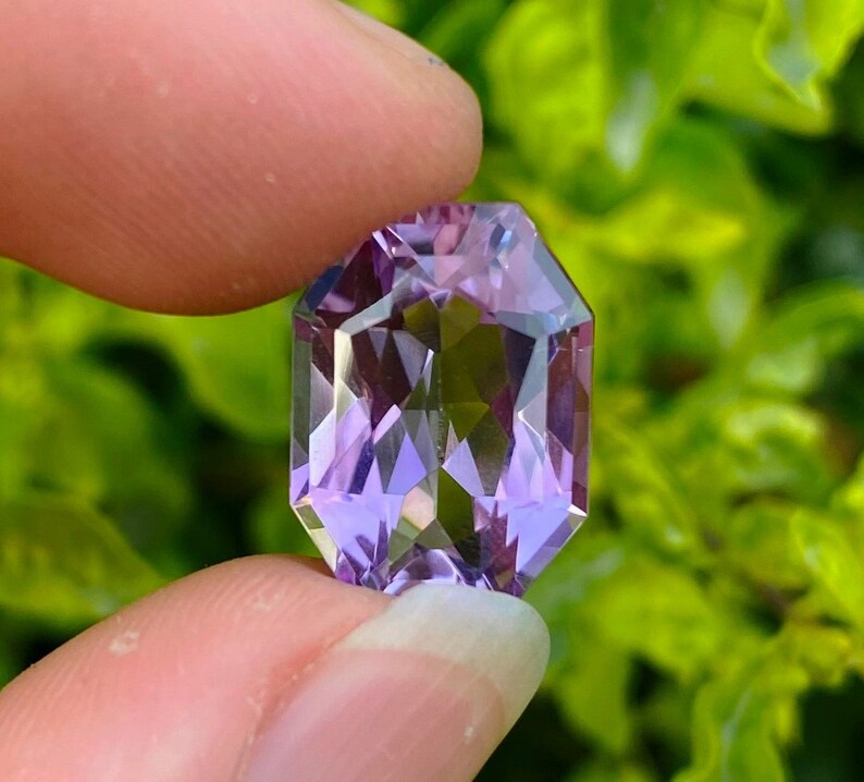 Peut inclure: Une grande gemme d'am&eacute;thyste facet&eacute;e de forme octogonale avec une teinte violette. La gemme est tenue dans une main, avec un fond de feuilles vertes.
