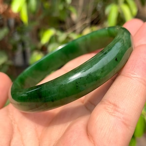 Puede incluir: Un brazalete de jade verde pulido. El brazalete liso y circular tiene un acabado brillante y un color verde oscuro intenso con sutiles variaciones. El brazalete de jade es una pieza de joyería clásica.