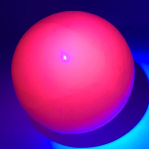 K&ouml;nnte beinhalten: Eine leuchtend pinkfarbene Kugel mit einem kleinen, hellen Licht in der Mitte, beleuchtet vor einem blauen Hintergrund.