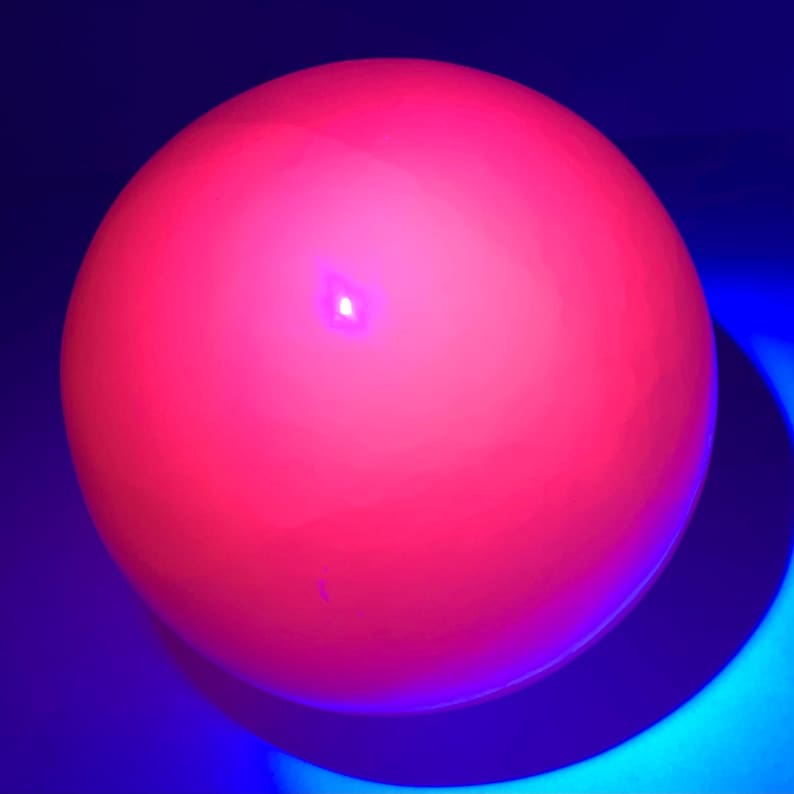 K&ouml;nnte beinhalten: Eine leuchtend pinkfarbene Kugel mit einem kleinen, hellen Licht in der Mitte, beleuchtet vor einem blauen Hintergrund.