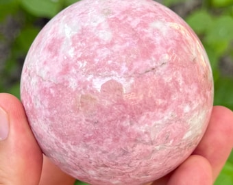 May include: A pink sphere with white and darker pink marbling. The stone ball has a smooth surface with natural variations in color and texture. The sphere is held in a hand, with a blurred green background.