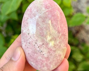 May include: A smooth, oval-shaped pink gemstone with white and tan marbling. The polished stone is held in a hand, with green foliage in the background. The stone's surface has small black speckles.