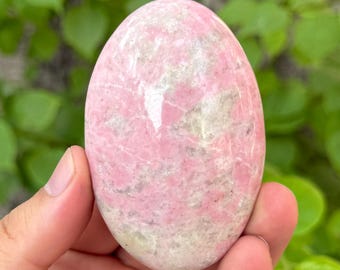 May include: A pink and white oval-shaped stone, held in a hand. The stone has a smooth, polished surface with a marbled appearance. The colors are a soft pink and off-white, with subtle variations in tone. The stone is a natural, organic shape.