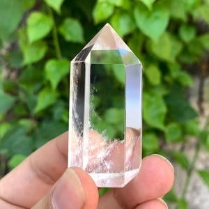 May include: A clear quartz crystal point with a hexagonal shape. The crystal is smooth and polished, with a slight cloudy appearance. It is held in a hand against a green background.