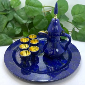 May include: A lapis lazuli coffee set featuring a dark blue coffee pot with a gold-colored spout and handle, a matching tray, and six small cups with gold-colored interiors. The set is displayed against a backdrop of green foliage.