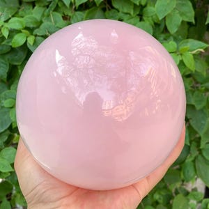 K&ouml;nnte beinhalten: Eine gro&szlig;e, polierte, rosafarbene Rosenquarzkugel. Die Kugel ist glatt und reflektierend, mit einer leichten Transluzenz.