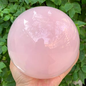 K&ouml;nnte beinhalten: Eine gro&szlig;e, glatte, rosafarbene Rosenquarzkugel. Die Kugel ist poliert und reflektiert Licht.