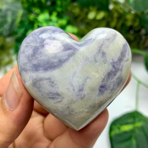 May include: A heart-shaped stone with a swirling pattern of lavender and white. The polished surface reflects light, highlighting the natural variations in the stone. The heart is held in a hand, with a green background.
