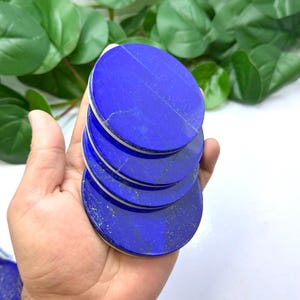 Op de afbeelding: Een stapel ronde, gepolijste lapis lazuli stenen. De stenen zijn diepblauw met subtiele marmering en een witte rand. De stenen worden in een hand gehouden, met groen gebladerte op de achtergrond.