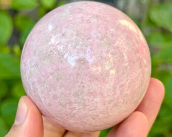 May include: A pink sphere with a mottled appearance, held in a hand. The sphere has a smooth, polished surface with variations in color, ranging from light to medium pink. The object is a decorative stone or crystal.