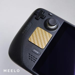 May include: A black handheld gaming device with a gold carbon fiber textured touchpad, a black joystick, and a black button labeled "STEAM". The device has the word "MEELO" printed on the side.