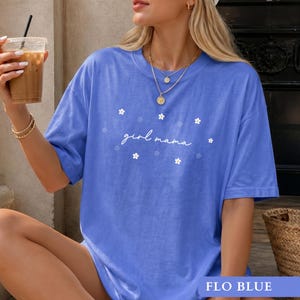 Op de afbeelding: Een korenblauw t-shirt met korte mouwen en de witte cursieve tekst "girl mama" in het midden op de borst, omringd door kleine witte bloemafbeeldingen. Het t-shirt heeft een ronde hals en een relaxte pasvorm van effen gekleurde stof.