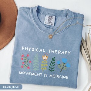 Op de afbeelding: Een blauw spijkerbroekkleurig t-shirt met de woorden "PHYSICAL THERAPY" en "MOVEMENT IS MEDICINE" op de voorkant. Het shirt heeft kleurrijke bloemenillustraties. Een gouden ketting ligt over het shirt.