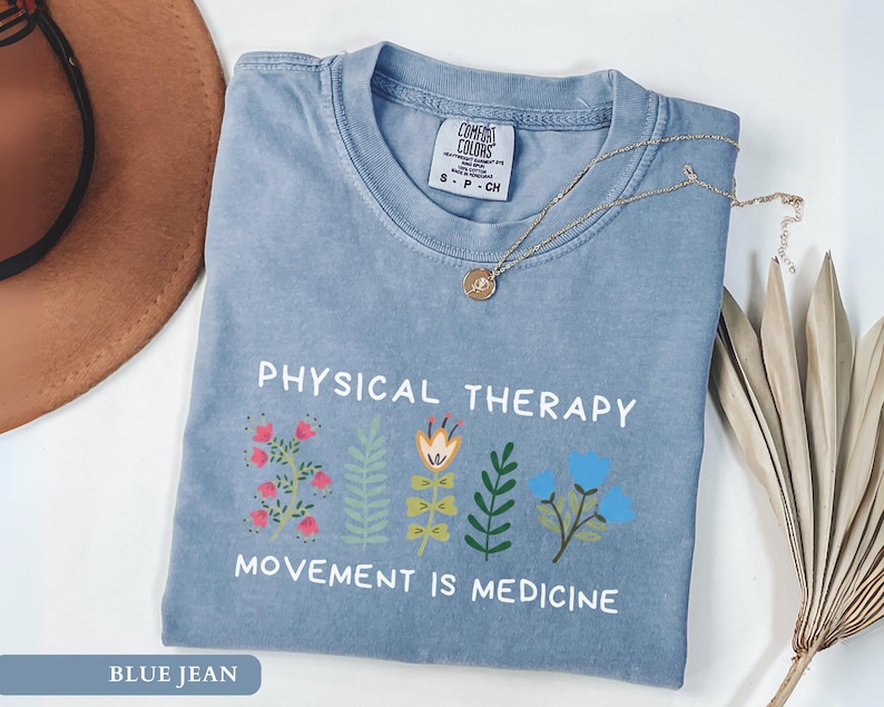 Op de afbeelding: Een blauw spijkerbroekkleurig t-shirt met de woorden "PHYSICAL THERAPY" en "MOVEMENT IS MEDICINE" op de voorkant. Het shirt heeft kleurrijke bloemenillustraties. Een gouden ketting ligt over het shirt.