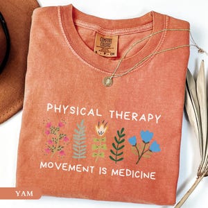 Op de afbeelding: Een koraalkleurig t-shirt met de woorden "PHYSICAL THERAPY" en "MOVEMENT IS MEDICINE" in het wit. Het shirt heeft bloemenillustraties in roze, blauw en groen. Een gouden ketting met een hanger ligt op het shirt.