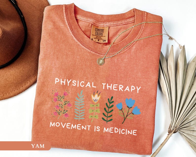 Op de afbeelding: Een koraalkleurig t-shirt met de woorden "PHYSICAL THERAPY" en "MOVEMENT IS MEDICINE" in het wit. Het shirt heeft bloemenillustraties in roze, blauw en groen. Een gouden ketting met een hanger ligt op het shirt.