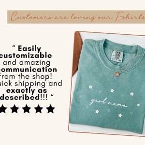 Op de afbeelding: Een saliegroen T-shirt met korte mouwen, voorzien van een klein wit bloemenpatroon en de tekst "girl mama" in wit schrift. Het shirt heeft een Comfort Colors-merklabel bij de hals en wordt getoond met een zilveren ketting.