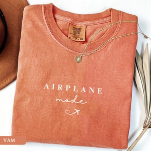 May include: A coral-coloured t-shirt with the text "AIRPLANE mode" in white lettering and a small airplane icon. The shirt is folded and laid on a white surface with a brown hat and a dried palm leaf.