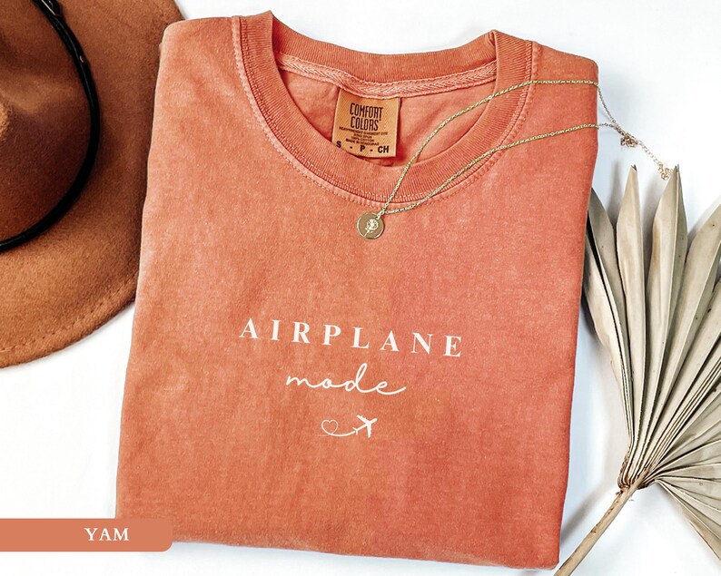 May include: A coral-coloured t-shirt with the text "AIRPLANE mode" in white lettering and a small airplane icon. The shirt is folded and laid on a white surface with a brown hat and a dried palm leaf.
