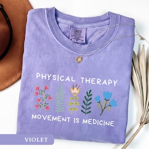 Op de afbeelding: Een violetkleurig T-shirt met de witte tekst "PHYSICAL THERAPY" en "MOVEMENT IS MEDICINE". Het heeft bloemenillustraties in roze, oranje, groen en blauw. Een gouden ketting ligt over het T-shirt.
