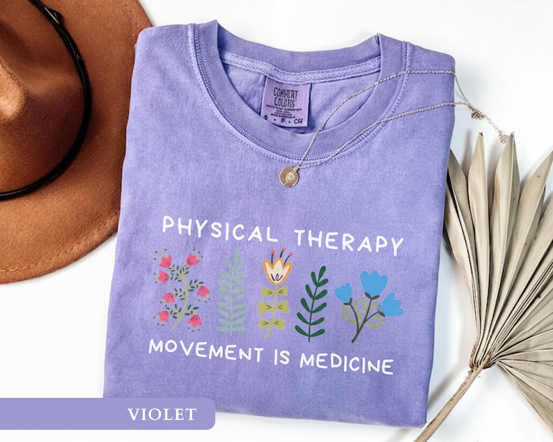 Op de afbeelding: Een violetkleurig T-shirt met de witte tekst "PHYSICAL THERAPY" en "MOVEMENT IS MEDICINE". Het heeft bloemenillustraties in roze, oranje, groen en blauw. Een gouden ketting ligt over het T-shirt.