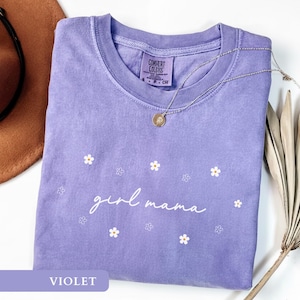 Op de afbeelding: Een violetkleurig T-shirt met korte mouwen en een ronde hals, voorzien van de witte tekst "girl mama" in cursief schrift, omringd door kleine witte en omlijnde bloemafbeeldingen. Het shirt heeft een Comfort Colors label in de nek.
