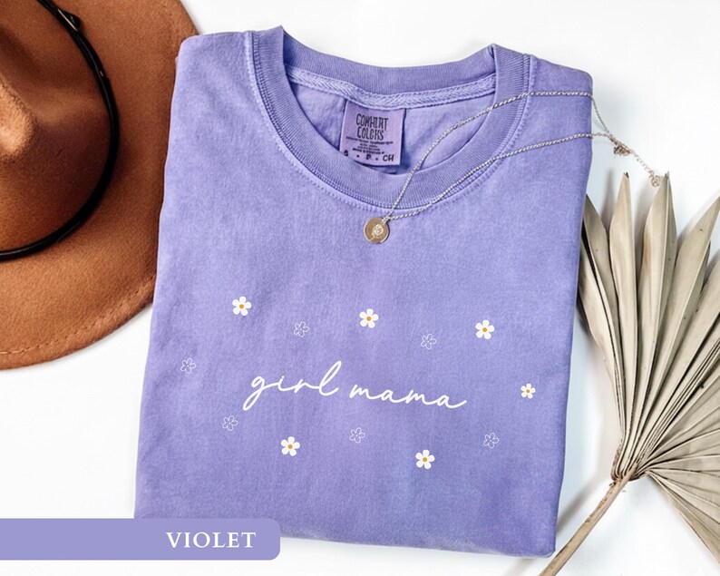 Op de afbeelding: Een violetkleurig T-shirt met korte mouwen en een ronde hals, voorzien van de witte tekst "girl mama" in cursief schrift, omringd door kleine witte en omlijnde bloemafbeeldingen. Het shirt heeft een Comfort Colors label in de nek.