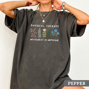 Op de afbeelding: Donkergrijs t-shirt met de witte tekst "PHYSICAL THERAPY" en "MOVEMENT IS MEDICINE". Het heeft een bloemenontwerp in roze, groen en blauw. Het woord "PEPPER" staat onderaan het shirt.