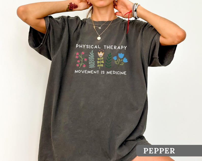 Op de afbeelding: Donkergrijs t-shirt met de witte tekst "PHYSICAL THERAPY" en "MOVEMENT IS MEDICINE". Het heeft een bloemenontwerp in roze, groen en blauw. Het woord "PEPPER" staat onderaan het shirt.