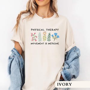 Op de afbeelding: Ivoor kleurig T-shirt met de tekst "PHYSICAL THERAPY" boven "MOVEMENT IS MEDICINE" en bloemen illustraties. Het shirt heeft een relaxte pasvorm, met een ronde hals en korte mouwen. Het model draagt een spijkerjasje.