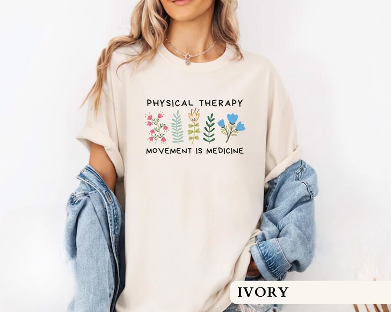 Op de afbeelding: Ivoor kleurig T-shirt met de tekst "PHYSICAL THERAPY" boven "MOVEMENT IS MEDICINE" en bloemen illustraties. Het shirt heeft een relaxte pasvorm, met een ronde hals en korte mouwen. Het model draagt een spijkerjasje.