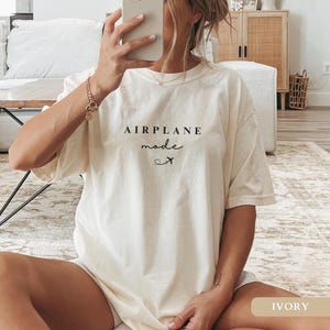 May include: An oversized white t-shirt with the text "AIRPLANE mode" and a small airplane icon. The shirt is being worn by a person sitting on a rug. The text "IVORY" is visible in the bottom right corner of the image.