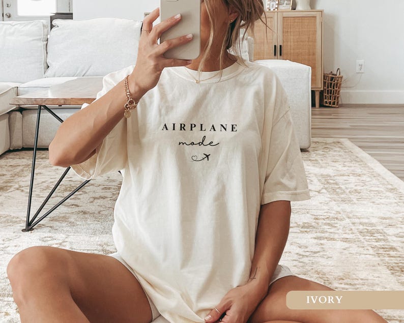 May include: An oversized white t-shirt with the text "AIRPLANE mode" and a small airplane icon. The shirt is being worn by a person sitting on a rug. The text "IVORY" is visible in the bottom right corner of the image.