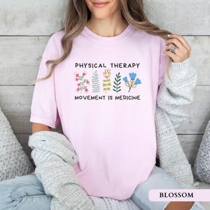Op de afbeelding: Een lichtroze t-shirt met de tekst "PHYSICAL THERAPY MOVEMENT IS MEDICINE" en bloemenillustraties. Het shirt is gemaakt van een zacht, comfortabel materiaal. Het woord "BLOSSOM" staat onderaan de afbeelding.