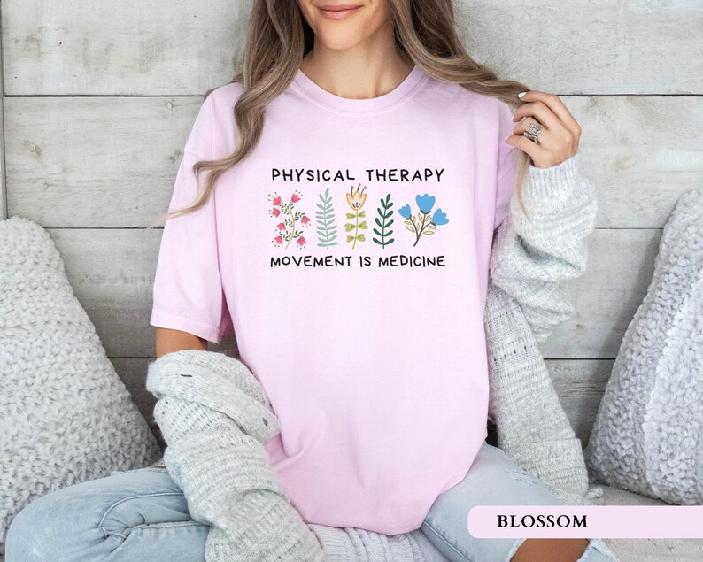 Op de afbeelding: Een lichtroze t-shirt met de tekst "PHYSICAL THERAPY MOVEMENT IS MEDICINE" en bloemenillustraties. Het shirt is gemaakt van een zacht, comfortabel materiaal. Het woord "BLOSSOM" staat onderaan de afbeelding.