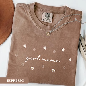 Op de afbeelding: Een bruin T-shirt met korte mouwen en een ronde hals, voorzien van de witte tekst "girl mama" in cursief schrift, omringd door kleine witte en omlijnde bloemillustraties. Het shirt heeft een Comfort Colors label in de kraag.