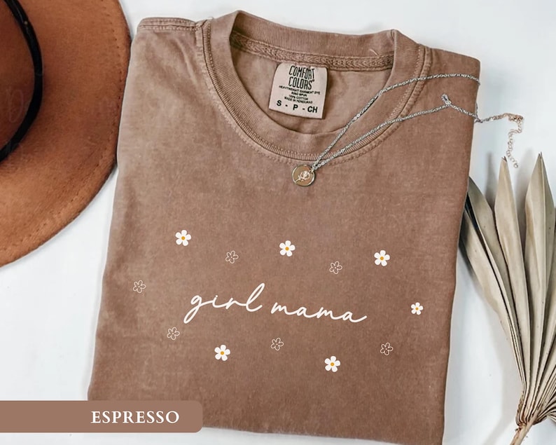 Op de afbeelding: Een bruin T-shirt met korte mouwen en een ronde hals, voorzien van de witte tekst "girl mama" in cursief schrift, omringd door kleine witte en omlijnde bloemillustraties. Het shirt heeft een Comfort Colors label in de kraag.
